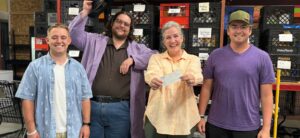 Smiling woman holding a $2500 check with three younger men standing on either side of her. They are standing in front of plastic crates filled with donated food.